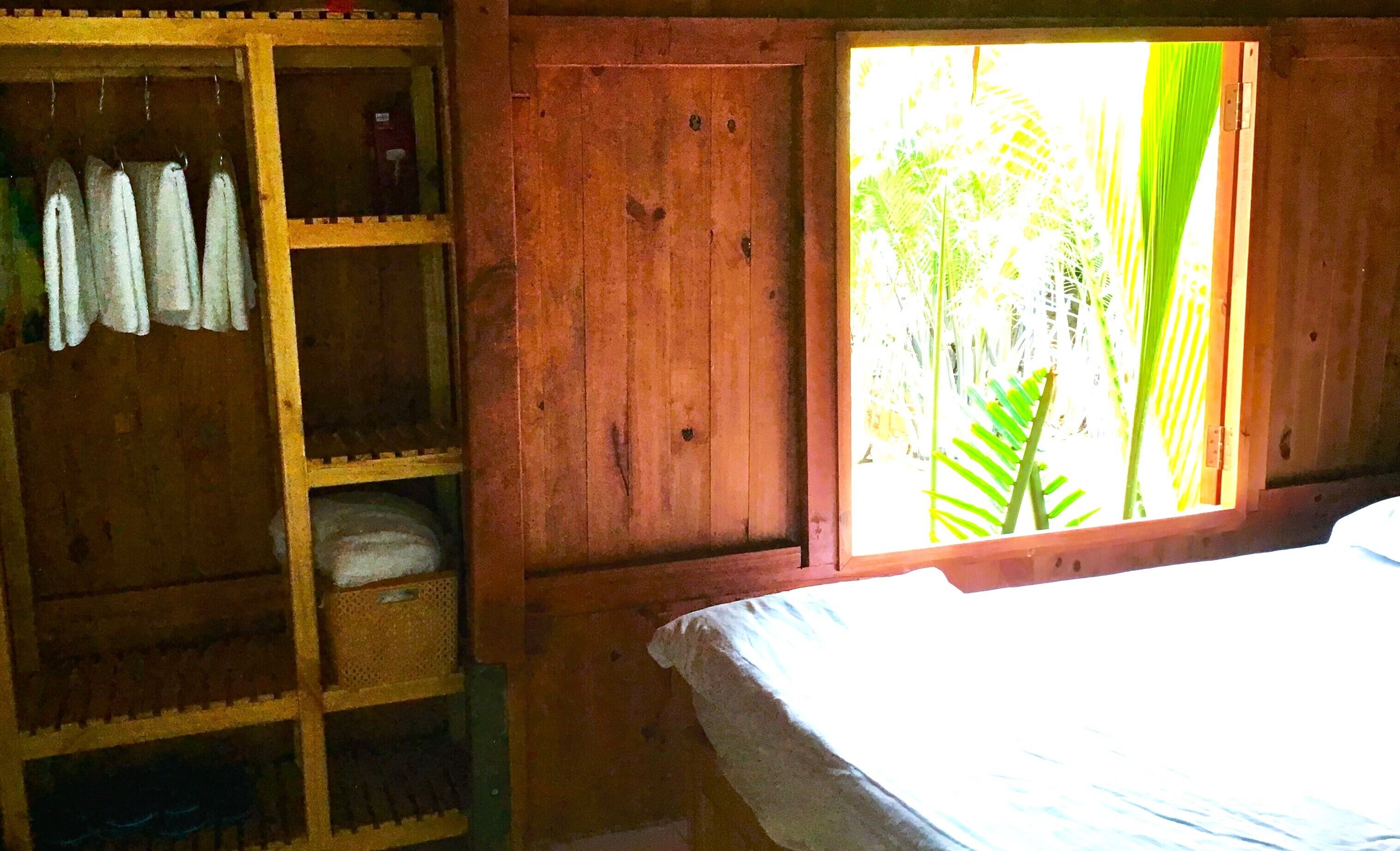 Una habitación de madera con una cama, un estante con ropa colgada y una ventana con vistas a la vegetación.