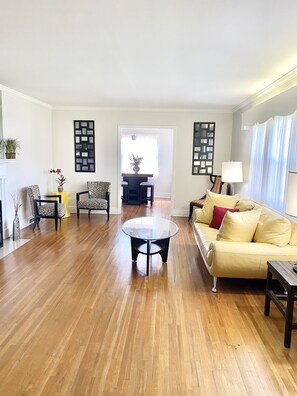Amplia sala de estar con pisos de madera, una mesa de centro redonda de vidrio, un sofá color beige y dos sillones. En el fondo, se observa un comedor con una mesa y sillas.