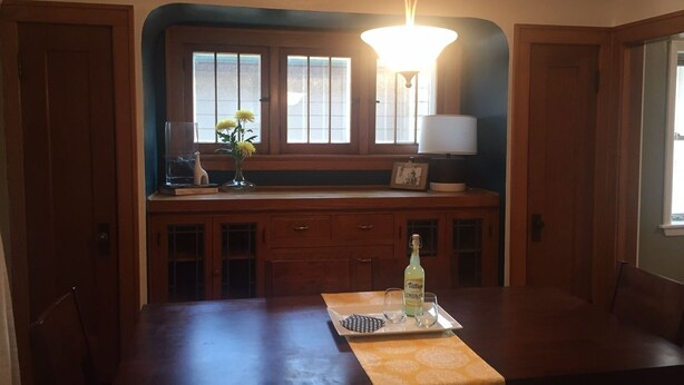 Un coin repas avec une table en bois, un lustre, une fenêtre avec des stores et un vase rempli de fleurs posé sur le rebord de la fenêtre.
