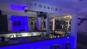 Zona de bar con luz azul neón, una barra con diferentes botellas y un área de asientos con taburetes.