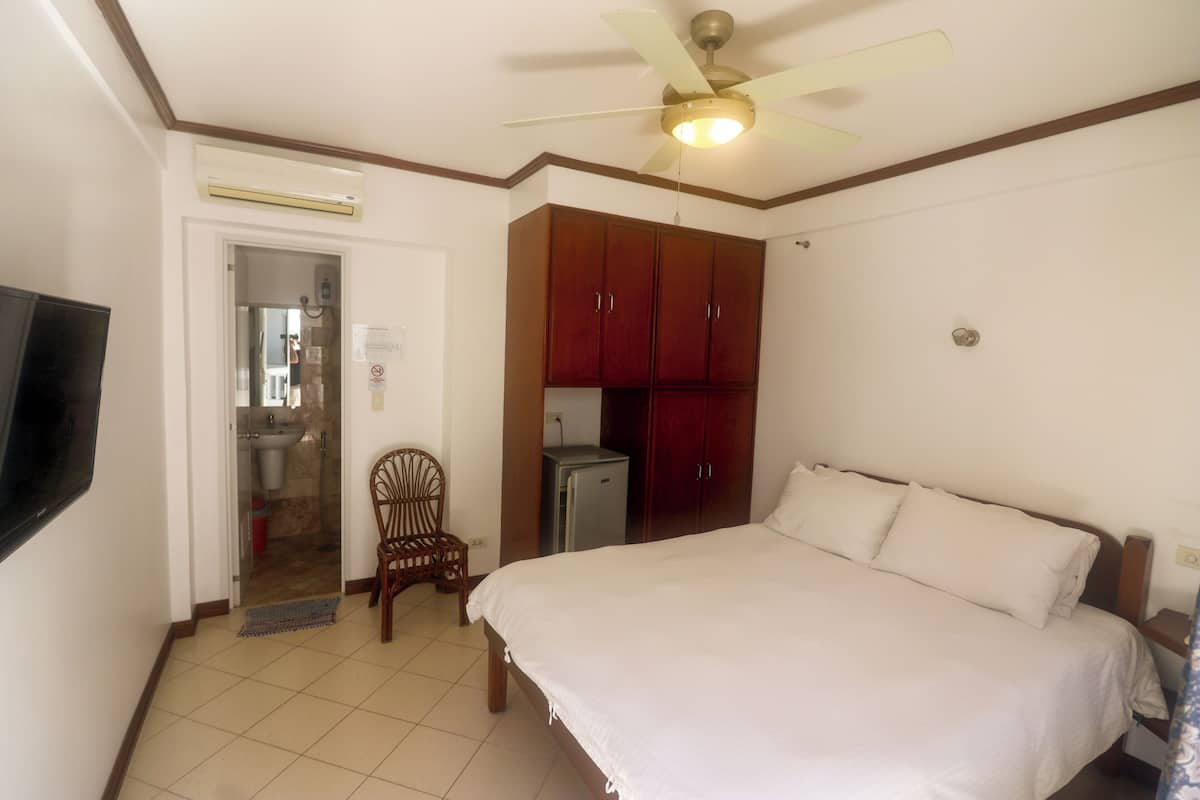 Une chambre comprenant un lit, un ventilateur de plafond, une télévision, une armoire et un petit coin cuisine.