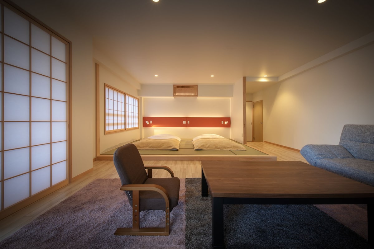 Ein modernes Hotelzimmer mit einem Bett auf Tatami-Matten, einem Holzstuhl, einer Couch und einem kleinen Tisch.