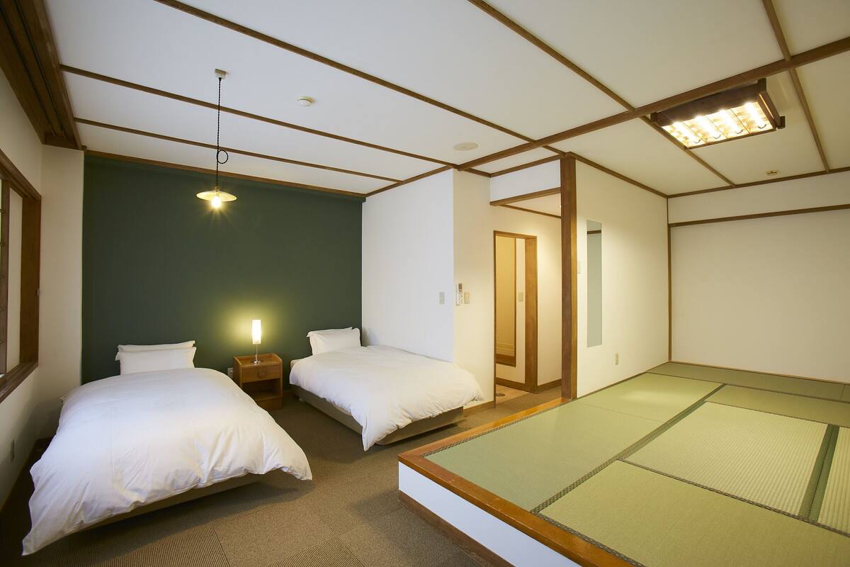 Ein Zimmer mit zwei Betten, einem kleinen Nachttisch mit Lampe, einer Zone mit Tatami-Matten und einem Deckenventilator.