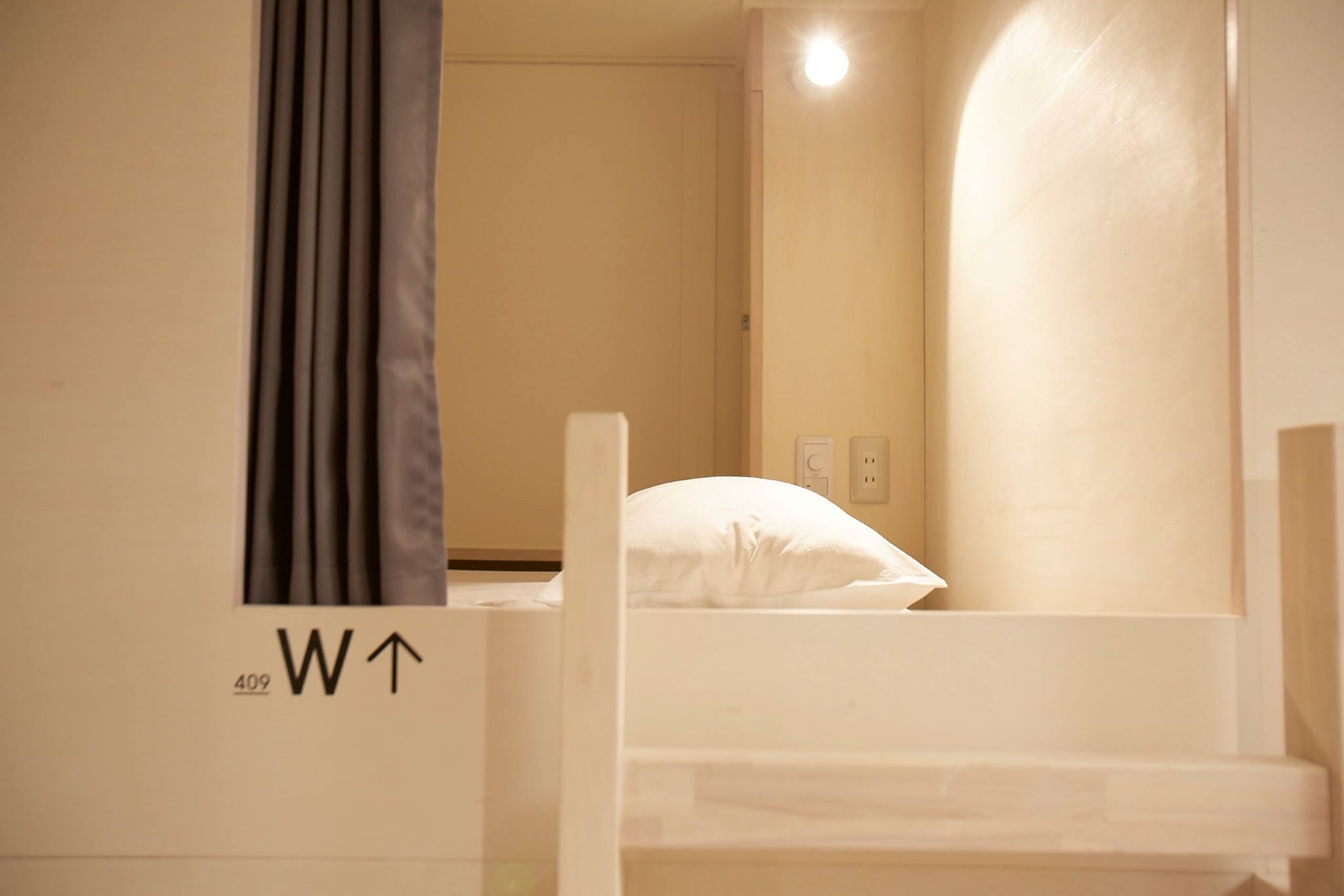 A single bed with a white pillow in a room with beige walls and a window with curtains.