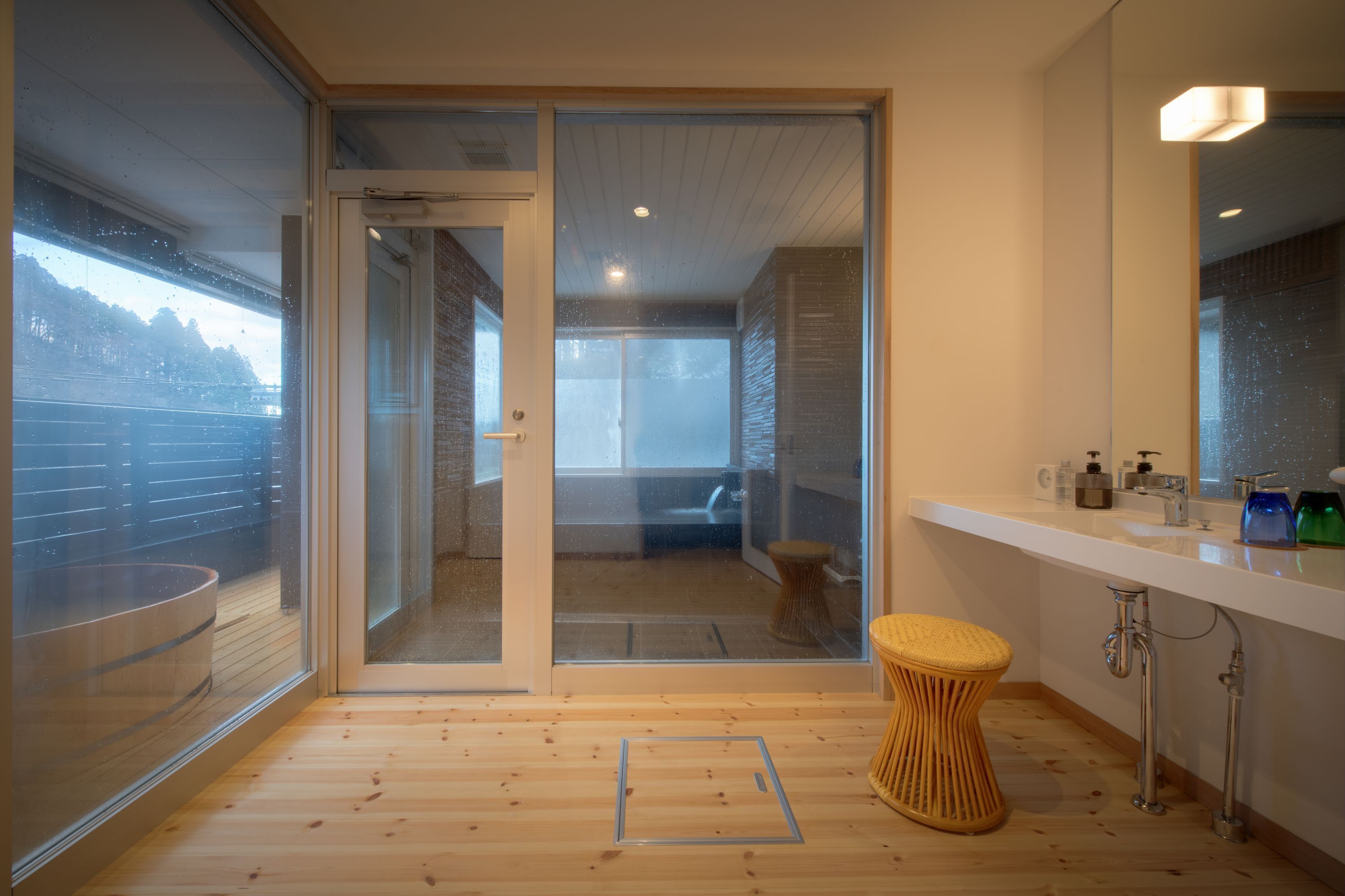 A modern bathroom with a glass door leading to a balcony, a wooden floor, a white sink, and a small stool.
