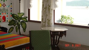 A room with a bed, a chair, a table, and a window with a view of a body of water and mountains. - Hotel Raj Niwas (Udaipur)