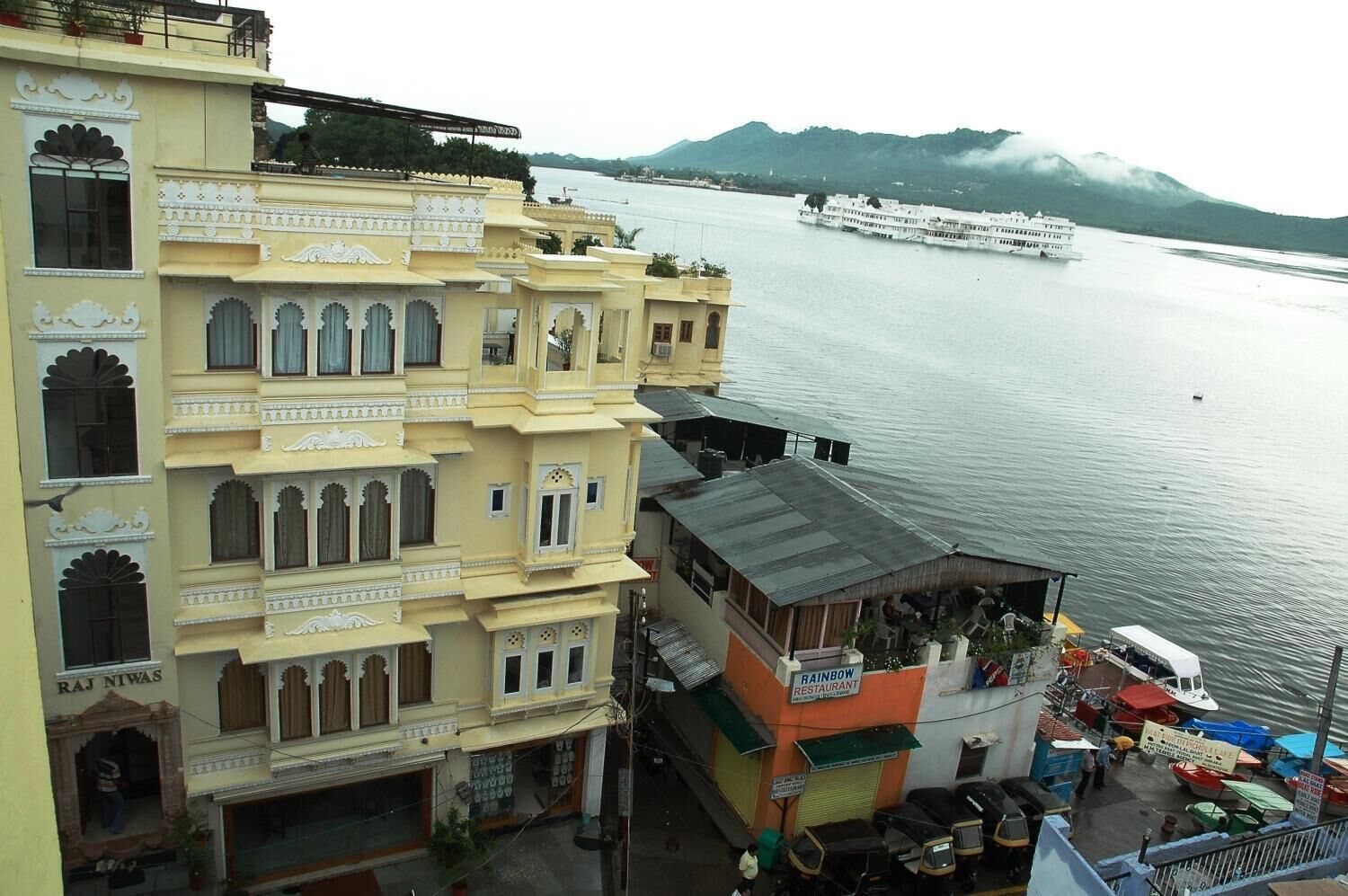 A multi-story building with traditional architectural elements, a view of a river with boats, and a mountainous backdrop.