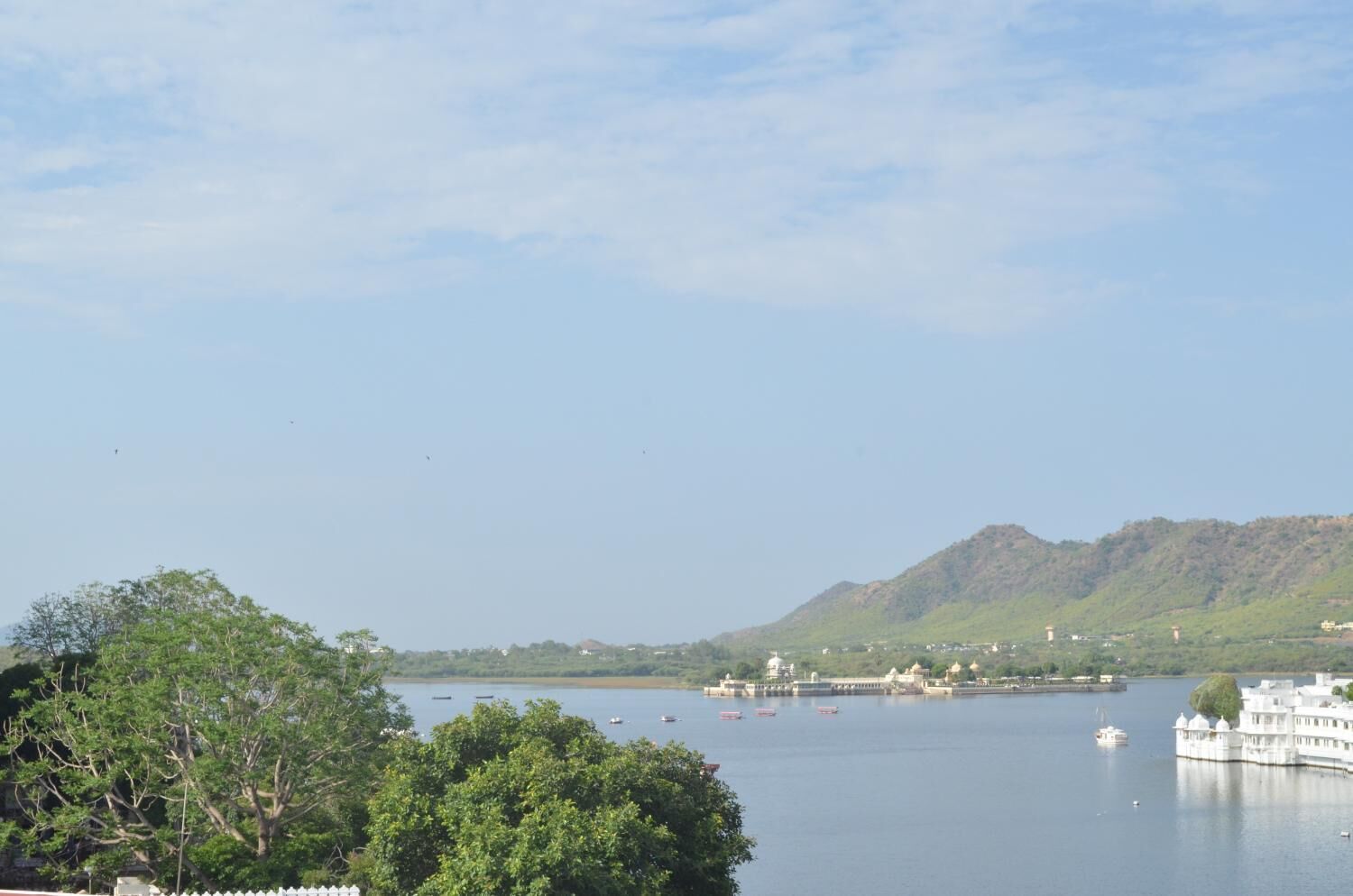 A calm lake with boats, a white building with domed structures, and a hill in the background.