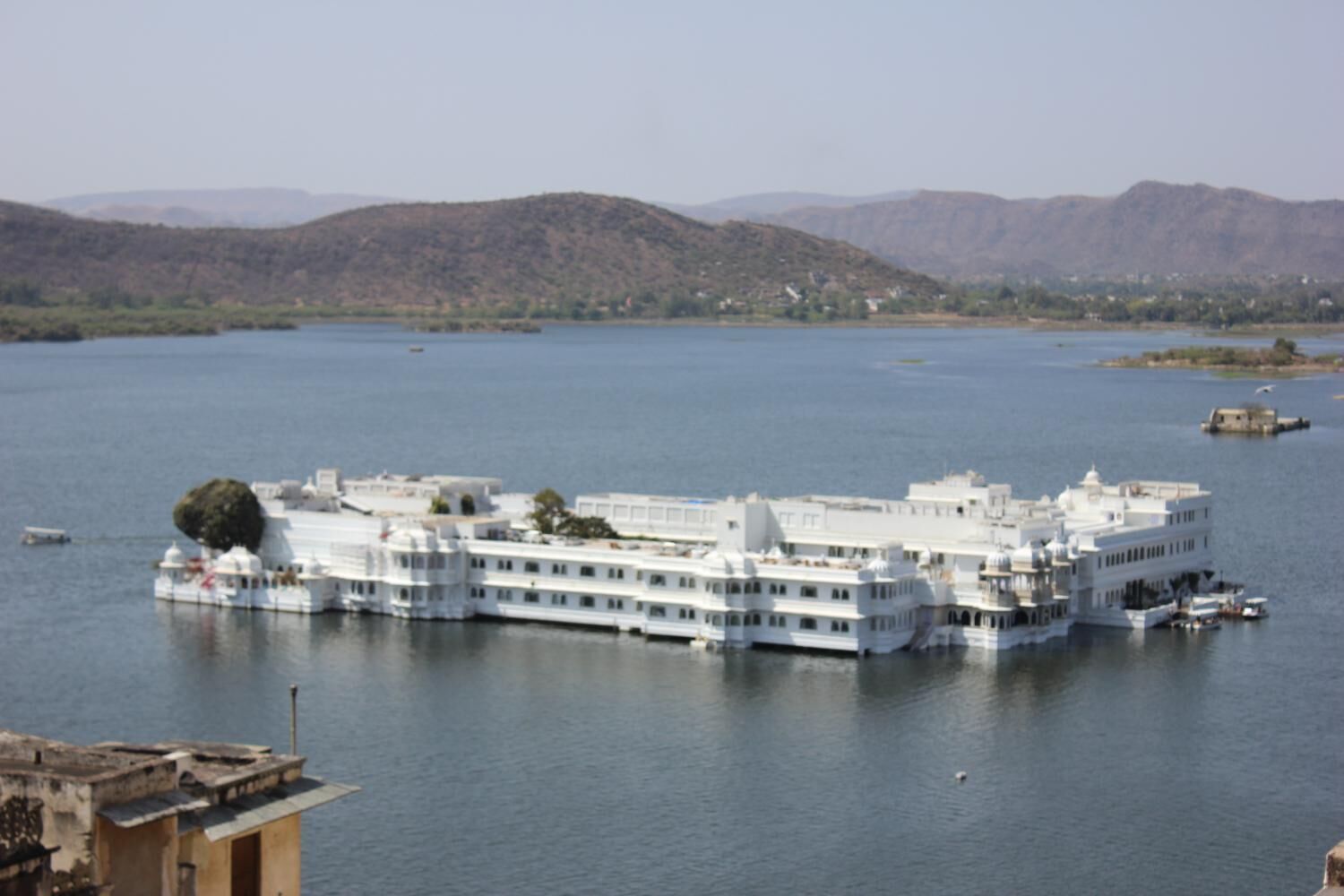 A large, white, multi-story building with a flat roof, situated on a lake surrounded by hills.