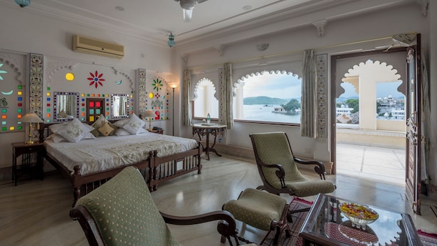 A spacious bedroom with a large bed, a sitting area with chairs, a small table, and a view of the city through ornate windows. - Hotel Raj Niwas (Udaipur)