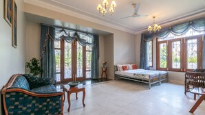 A spacious room with a large bed, a sofa, and a dining table. There are multiple windows with curtains, a chandelier, and a ceiling fan.