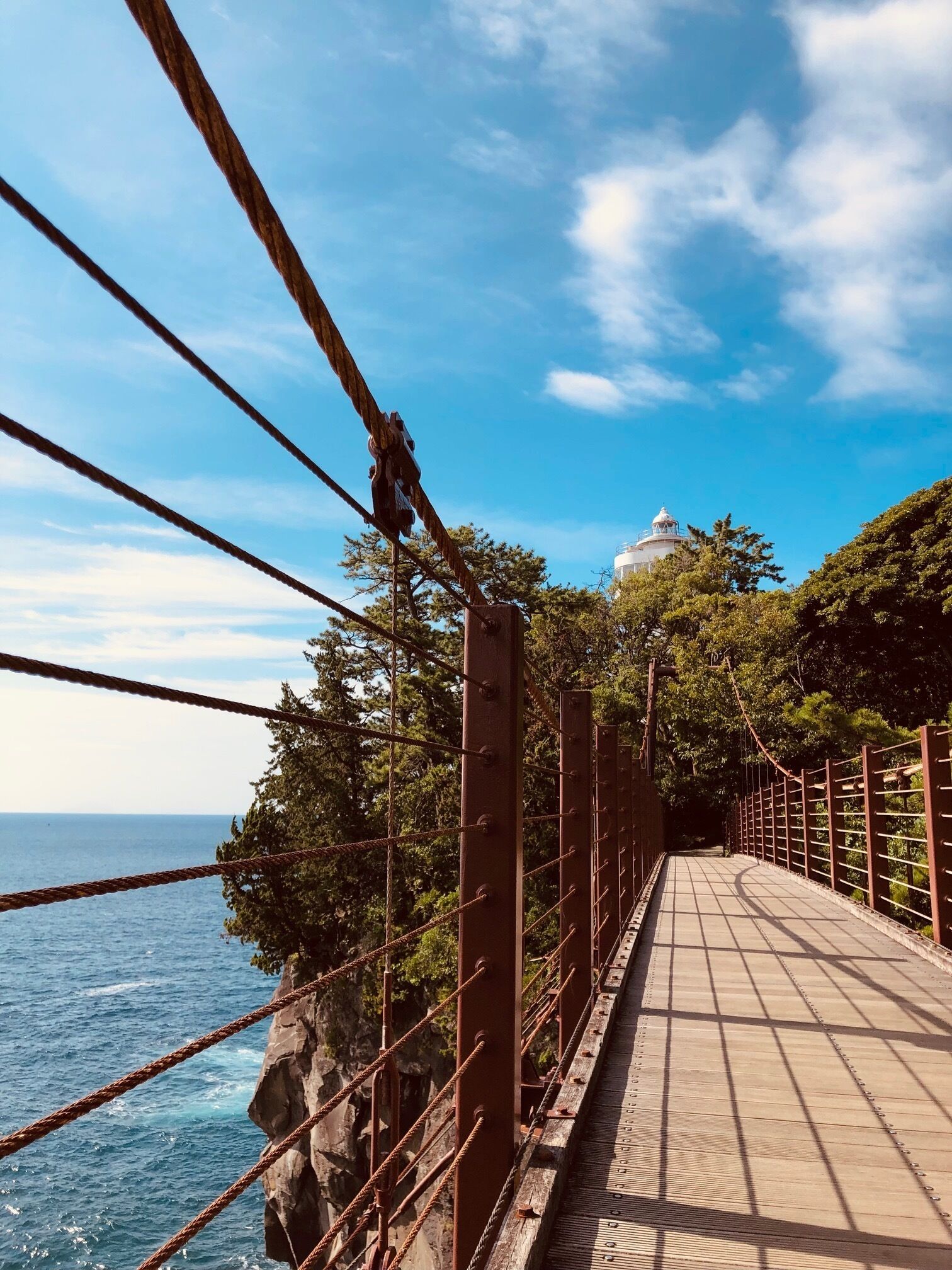 A suspension bridge with metal railings and a wooden walkway, overlooking a body of water and a white dome structure in the distance.