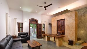 A spacious living room with a black leather sofa, a wooden coffee table, and a dining table. There is a ceiling fan, a fireplace, and a large window with curtains.