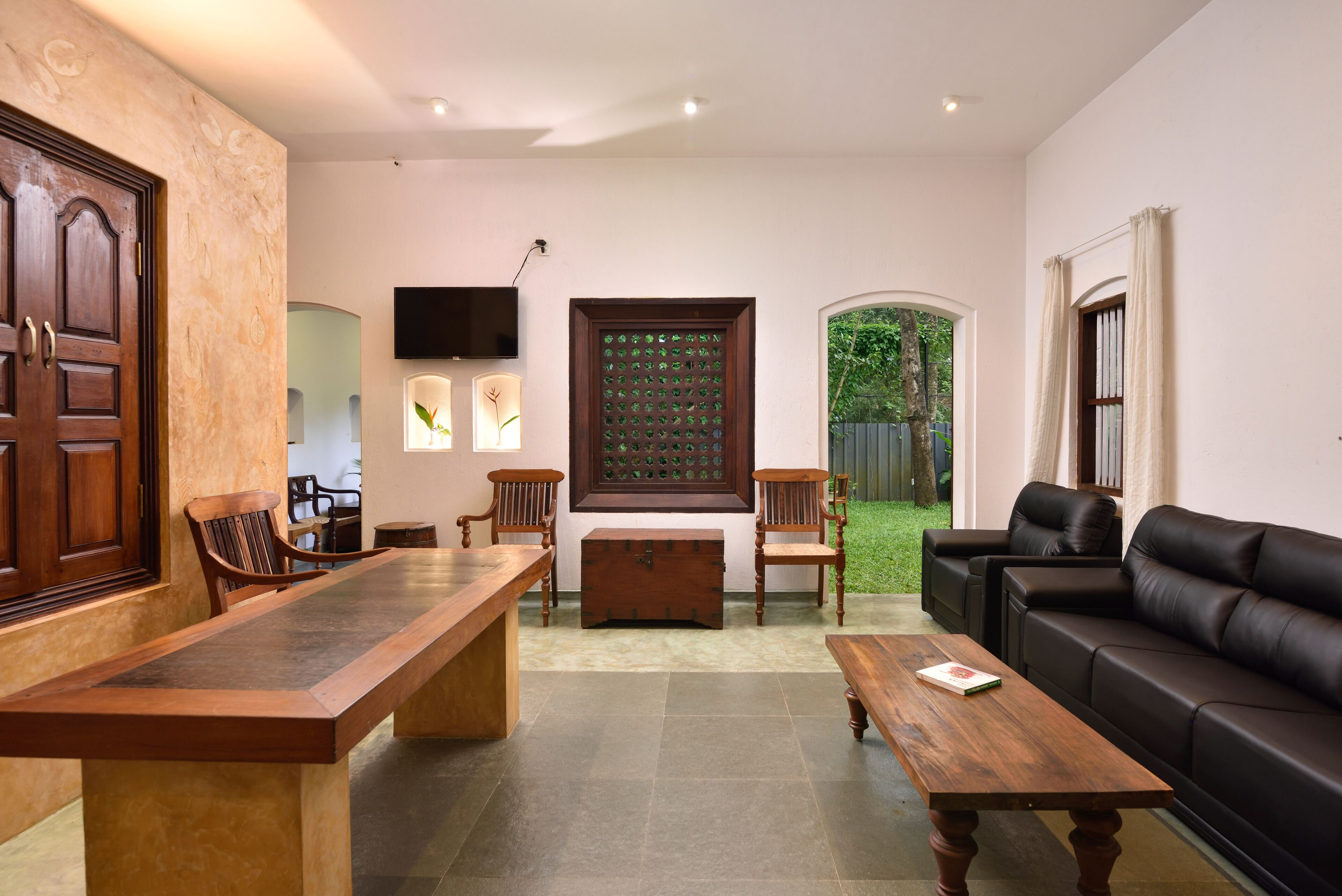 A spacious living room with a large wooden table, a black leather sofa, and a wooden bench. There is a television mounted on the wall and a window with a view of the outdoors.