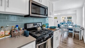 A modern kitchen with white cabinets, a stainless steel stove, and a microwave. There is a dining area with a blue sofa and a table.