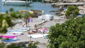 Una playa con un barco amarrado cerca de la orilla, un banco de madera y una pila de sillas blancas de plástico.
