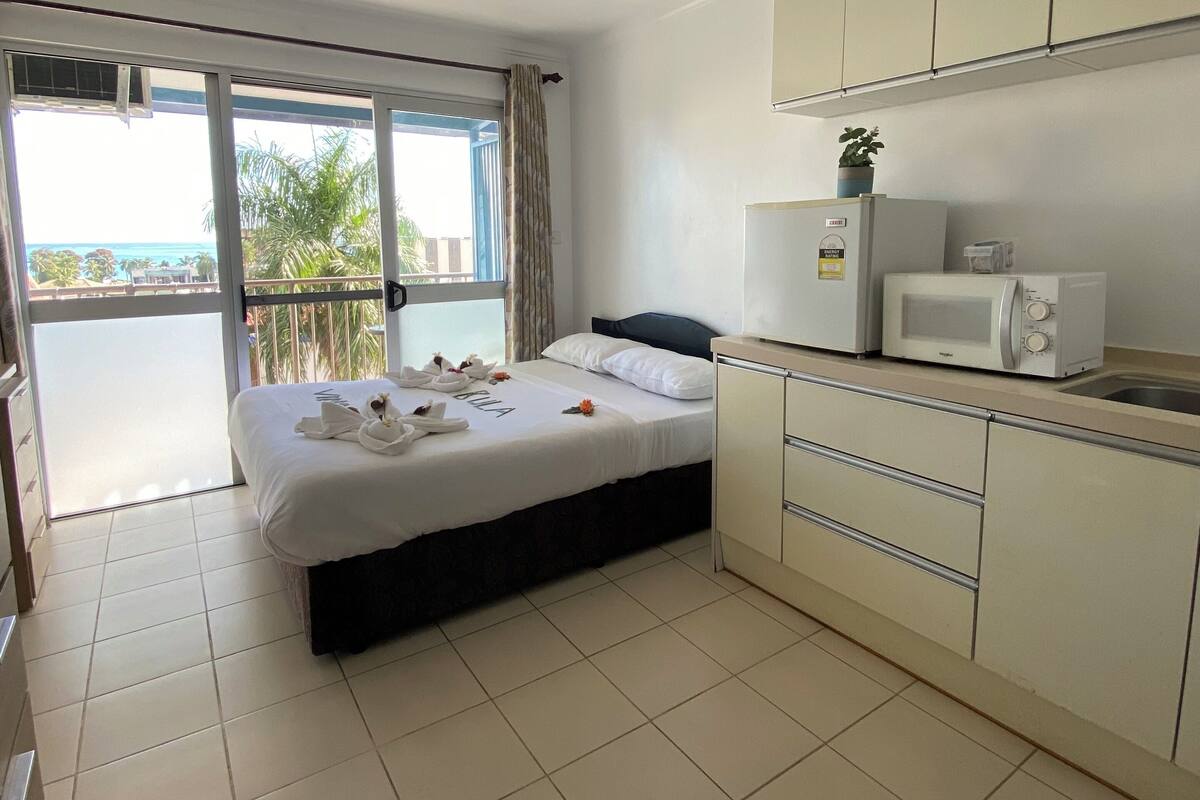 A hotel room with a bed, a kitchenette, and a balcony with a view of the ocean.