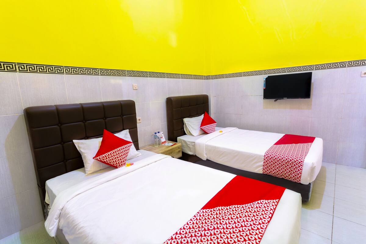 A hotel room with two beds, each with a red and white bedspread, a small bedside table with a bottle and a glass, and a flat-screen TV mounted on the wall.