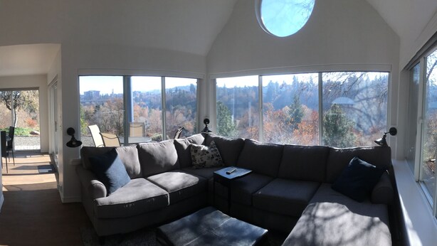 A cozy living room with a sectional sofa, a coffee table, and a TV mounted on the wall. Large windows offer a view of trees and a distant cityscape. - Kendall Yards Home - Stunning views, amazing downtown location (Spokane)