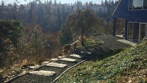 A house with a stone pathway leading to a deck, surrounded by trees and a view of distant buildings. - Kendall Yards Home - Stunning views, amazing downtown location (Spokane)