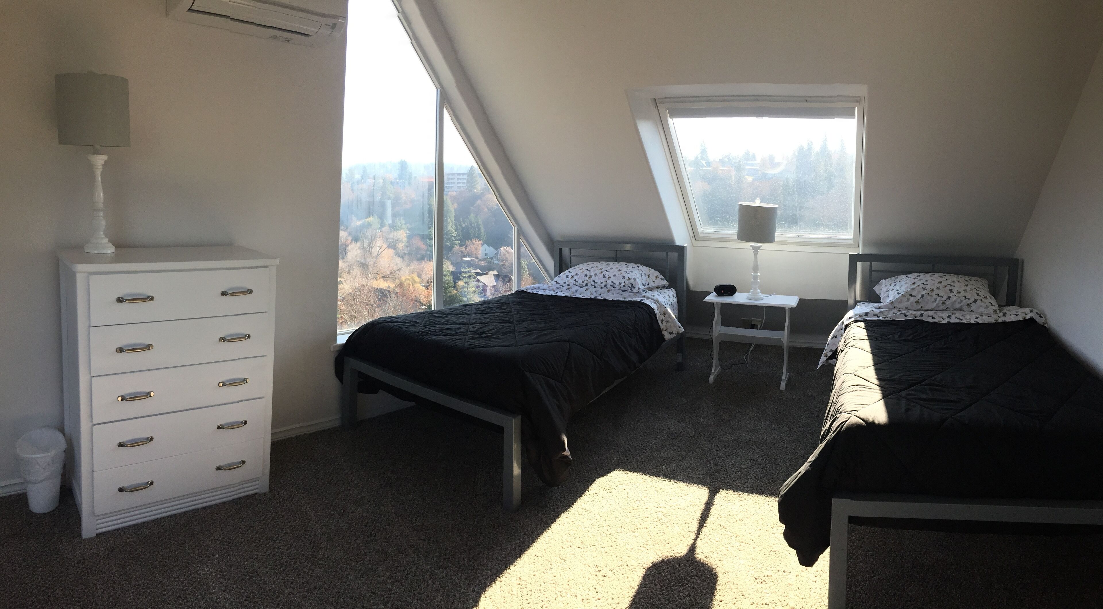 Ein Schlafzimmer mit zwei Betten, einem Schminktisch, einer Lampe und einem Fenster mit Blick auf Bäume.