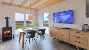 A modern kitchen with a wood-burning stove, a dining table with chairs, a TV mounted on the wall, and a view of the outdoors through large windows.