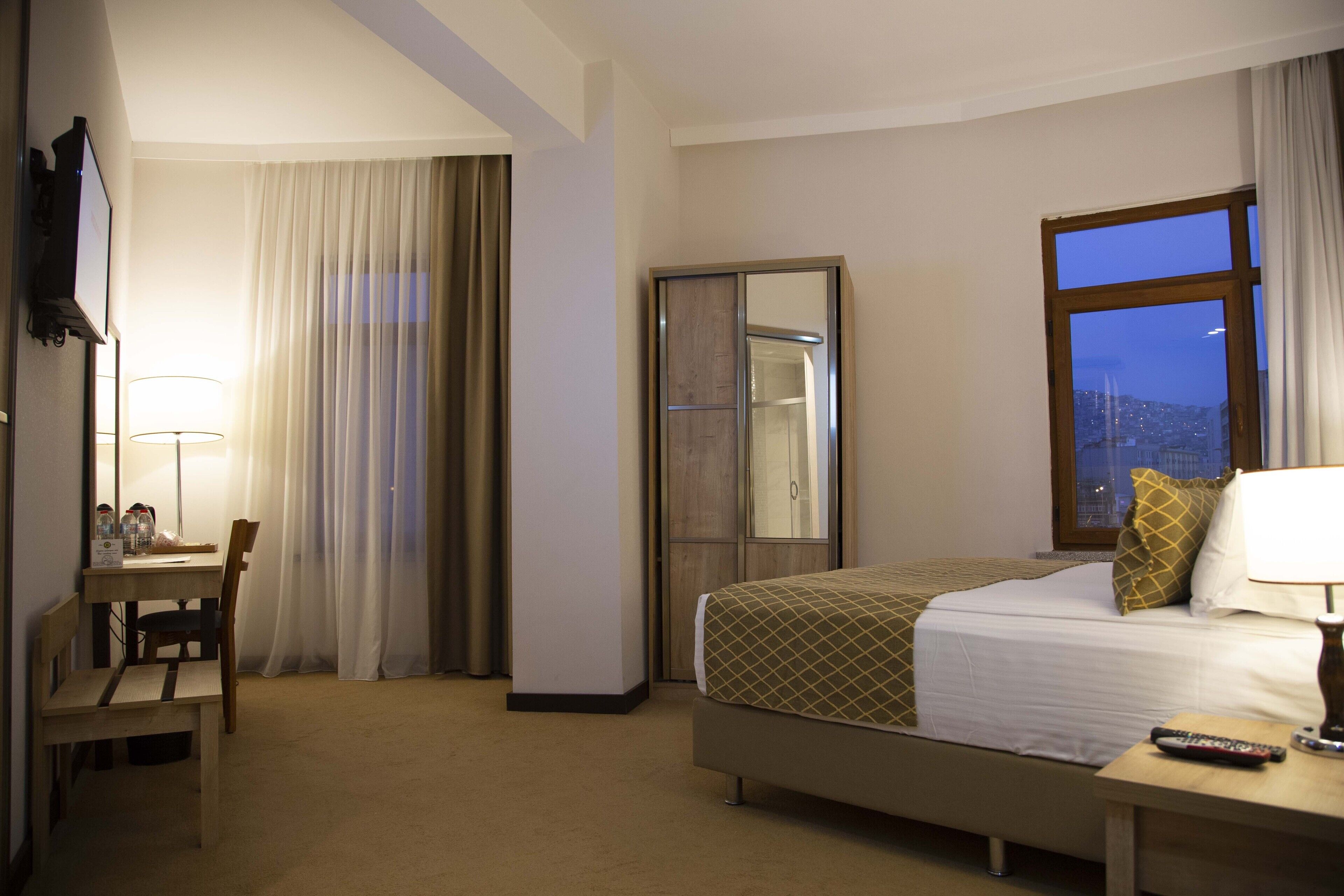 Habitación de hotel con cama, escritorio con lámpara, armario y ventana con vista a la ciudad.