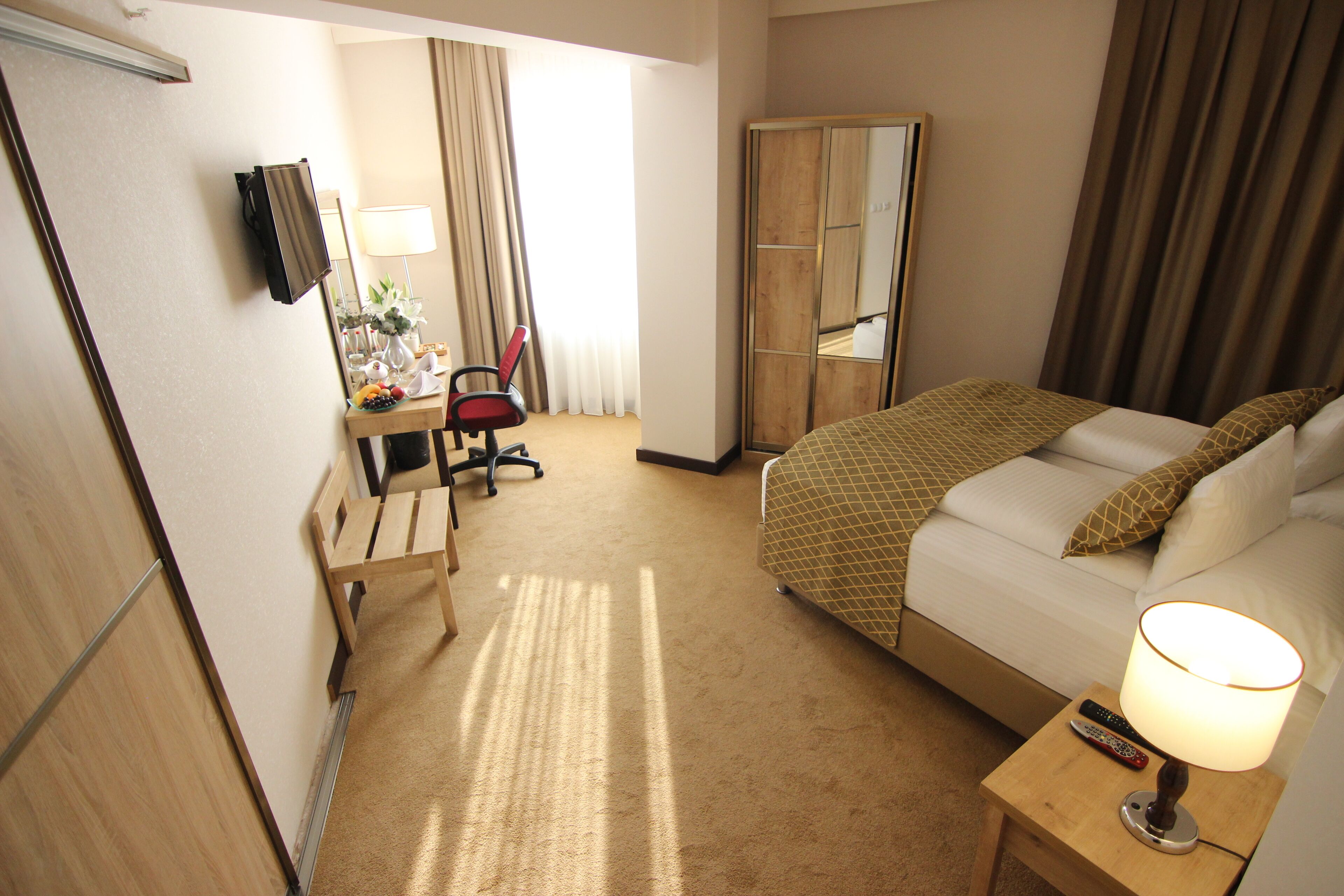 Una habitación de hotel con una cama, un escritorio con una silla, un espejo, una lámpara y un televisor en la pared.
