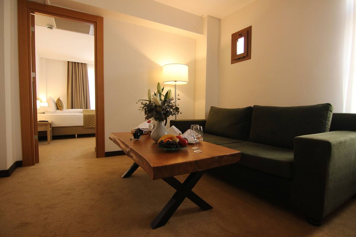 A hotel room with a wooden table, a green sofa, a lamp, and a bed with a view.