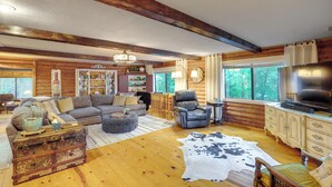 Interno di una baita in legno con soggiorno, divano, poltrona, poggiapiedi e un baule rustico.