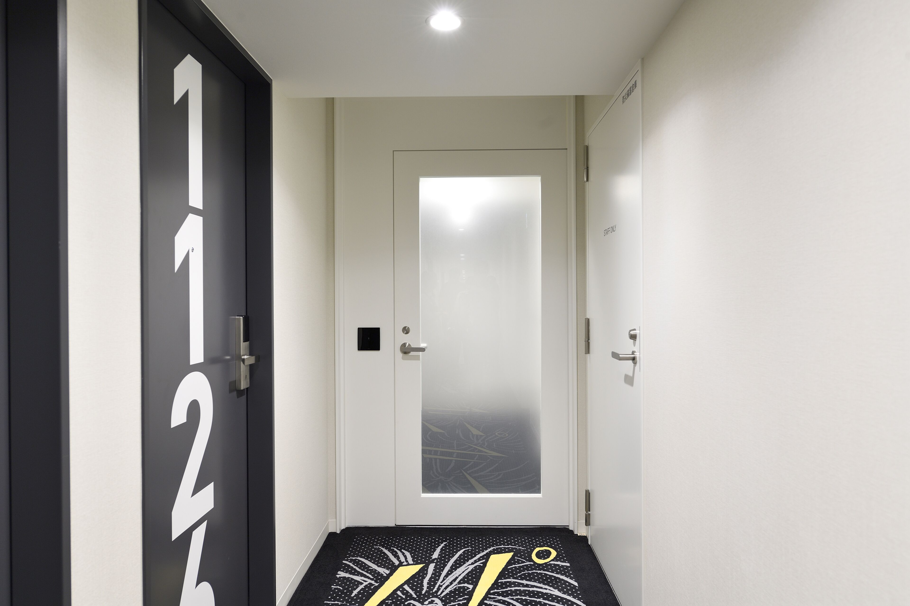 Um corredor estreito com uma porta de vidro fosco, uma porta preta com números brancos e um tapete de entrada com padrão preto e branco.