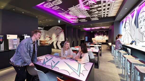 Un interior moderno con una mesa central rodeada de sillas, una zona de bar con taburetes y un techo con ilustraciones de estilo cómic iluminadas.