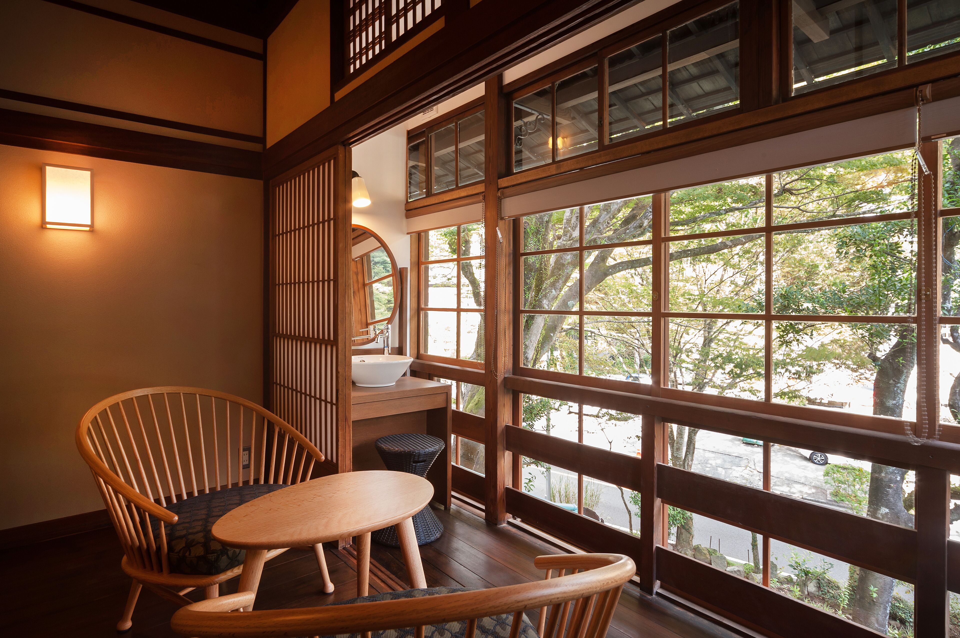 A traditional Japanese-style room with tatami flooring, a low wooden table, a cushioned chair, a washbasin, and a large window offering a view of trees and a street.
