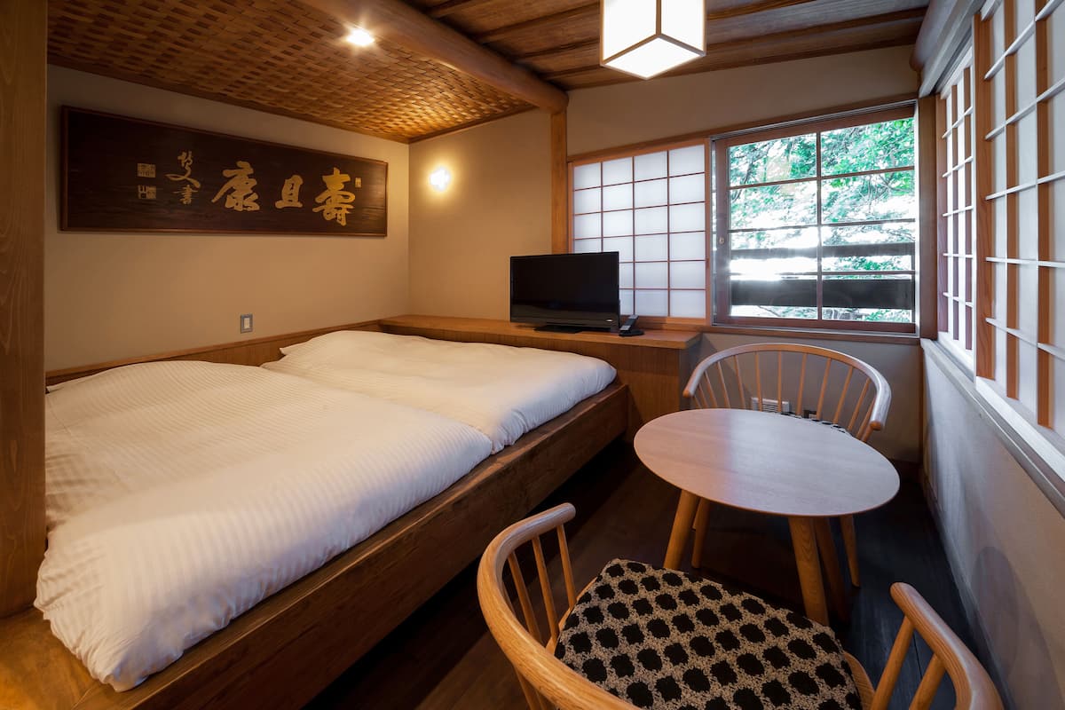 A traditional Japanese-style room with a tatami mat bed, a television, a small table, and a window with a view of greenery.