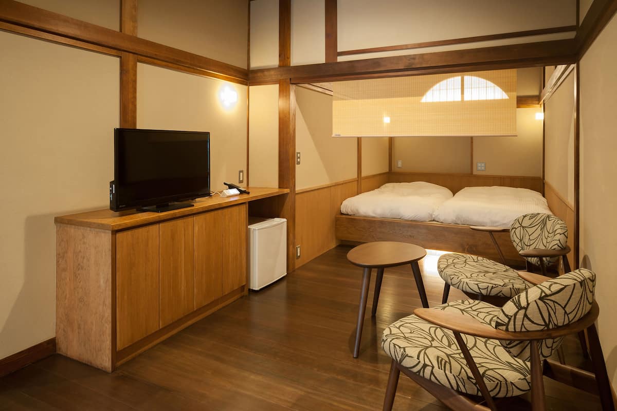 A compact room with a bed, a small desk with a television, a refrigerator, and a seating area with patterned chairs.