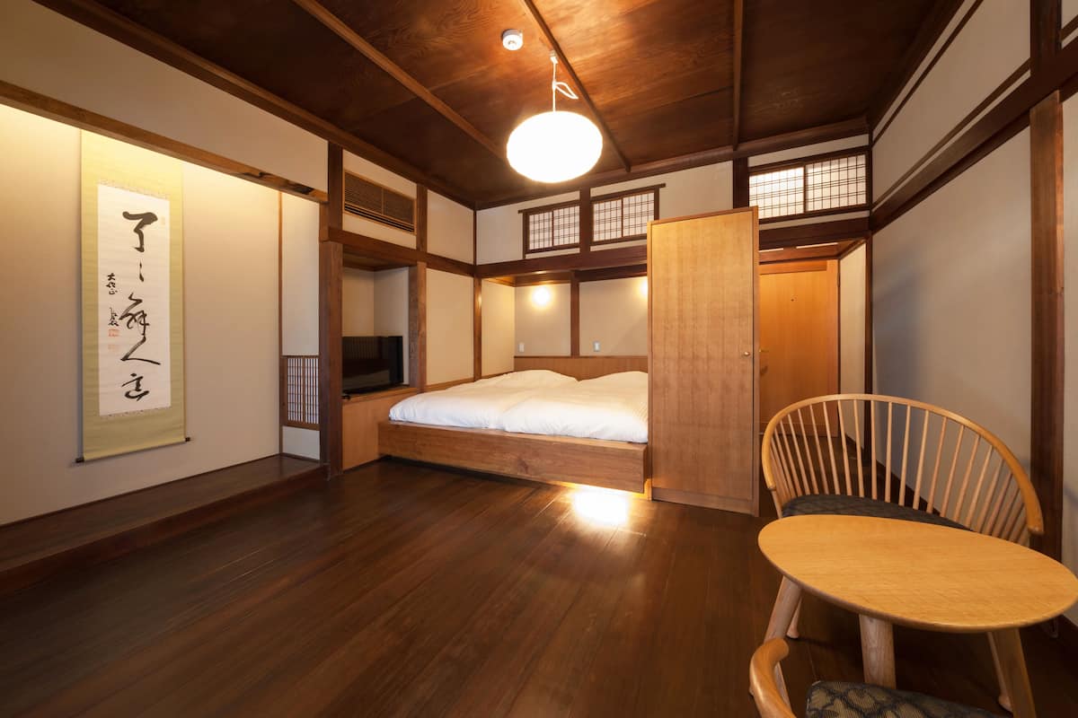 A traditional Japanese-style room with tatami flooring, a low wooden bed, a wooden chair, and a hanging shoji screen.
