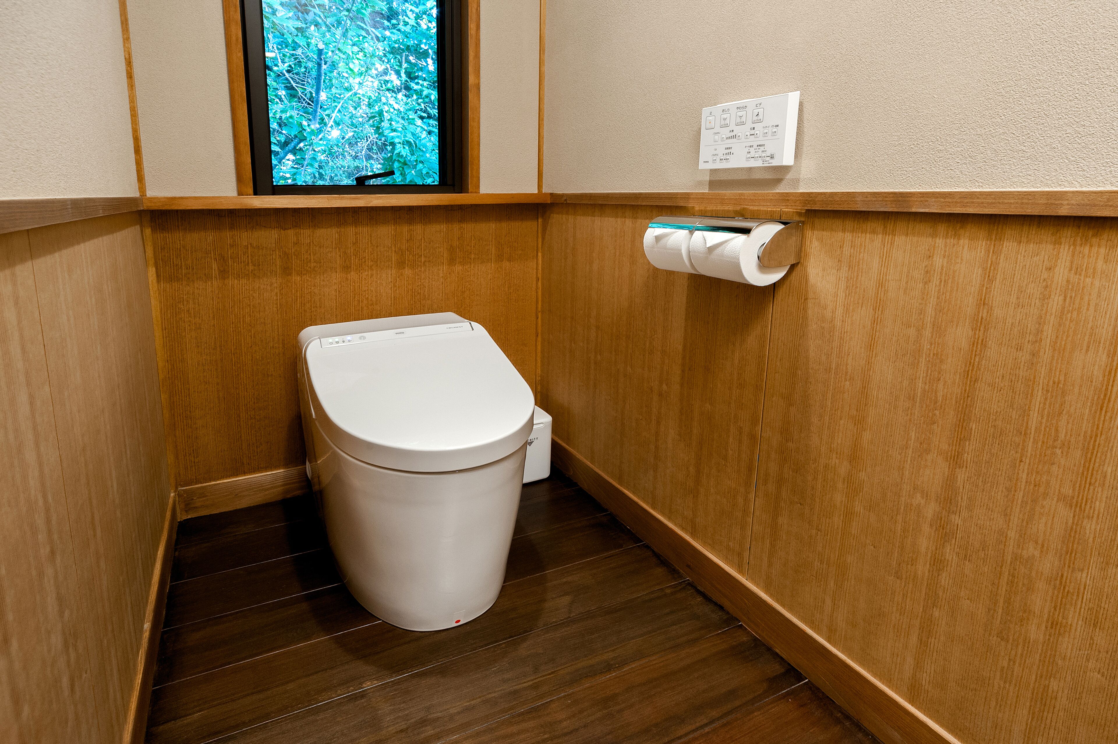 A small, compact bathroom with a wooden floor, a white toilet, and a window with a view of greenery.
