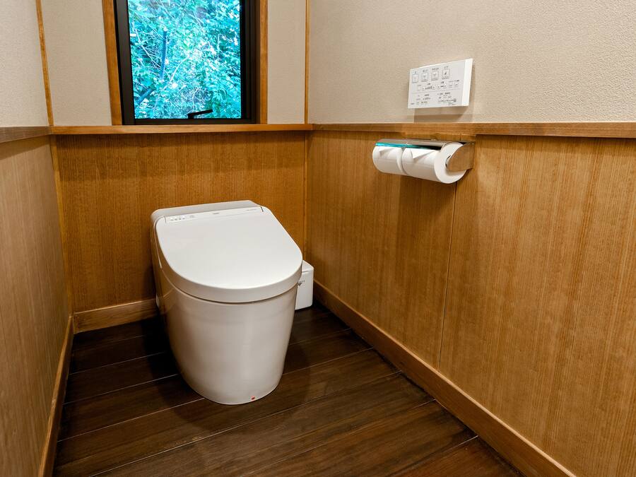 A small, compact bathroom with a wooden floor, a white toilet, and a window with a view of greenery.