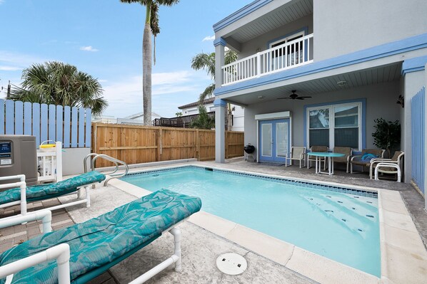 A swimming pool with lounge chairs, a wooden fence, and a covered patio area with outdoor furniture.