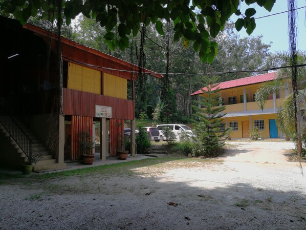 A two-story building with a red roof and yellow walls, surrounded by greenery and a gravel driveway.