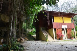Un bâtiment rustique avec un escalier menant à une porte jaune, entouré de verdure et bordé d’une grotte rocheuse.