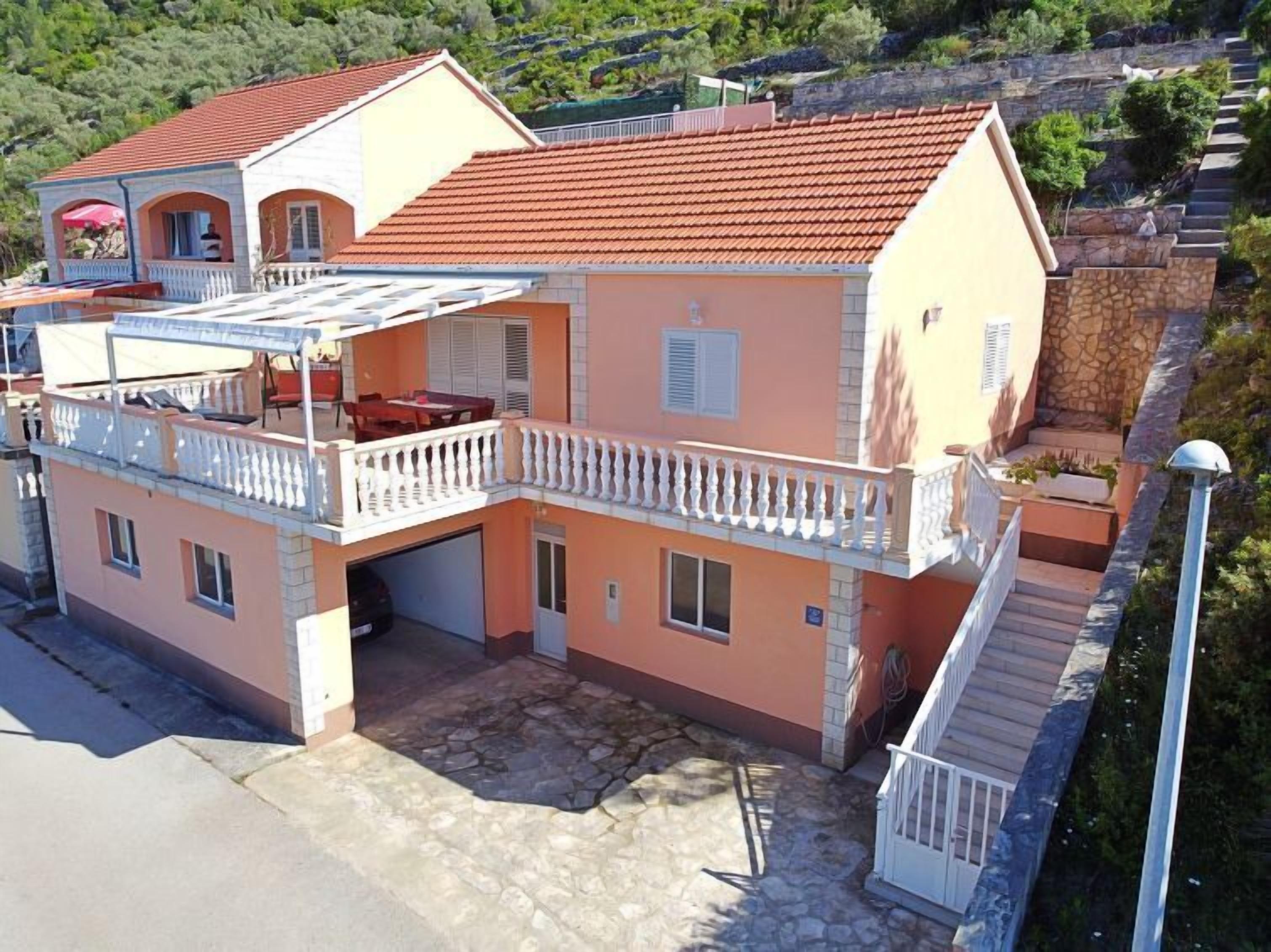 A two-story house with a red-tiled roof, a balcony with a white balustrade, and a stone-paved driveway.