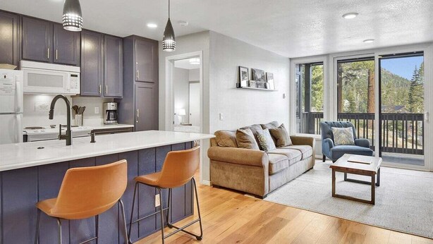 A modern kitchen with dark cabinets, a white countertop, and pendant lighting. There is a bar area with two stools and a sofa in the living area with a view of the outdoors. - Crestview 55: Pet Friendly, Close to The Village (Mammoth Lakes)