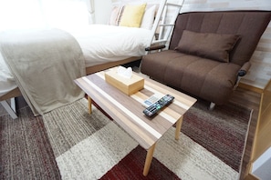 A hotel room with a bed, a brown sofa, a wooden table with a tissue box and remote control, and a striped rug. - Apartment Y Hakuyu Motomachi Namba (Osaka)