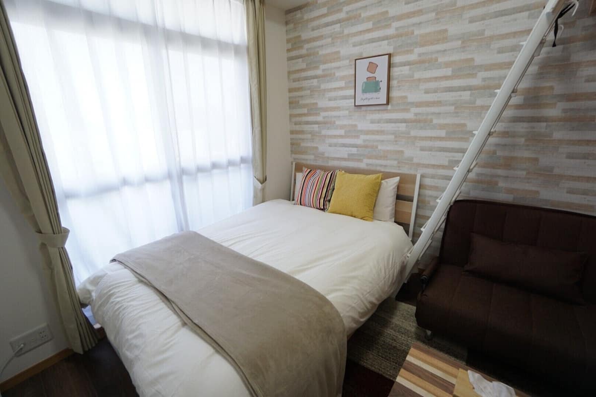 A bedroom with a bed, a brown sofa, a window with sheer curtains, and a wall-mounted picture.