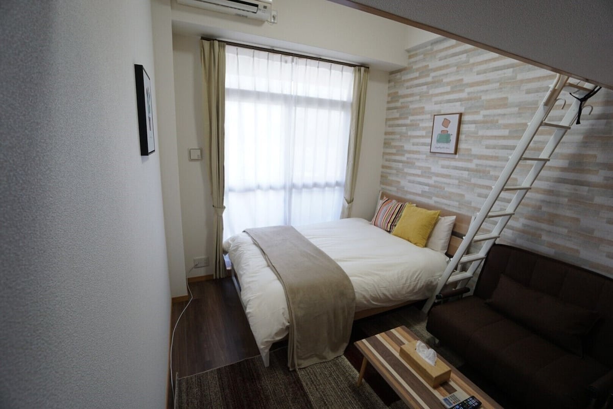 A bedroom with a bed, a bedside table, a brown sofa, a window with curtains, a wall-mounted air conditioner, and a wooden ladder.