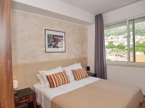 A hotel room with a bed, a nightstand, a framed picture on the wall, and a window with a view of a cityscape. - Apartments Xandy (Dubrovnik)
