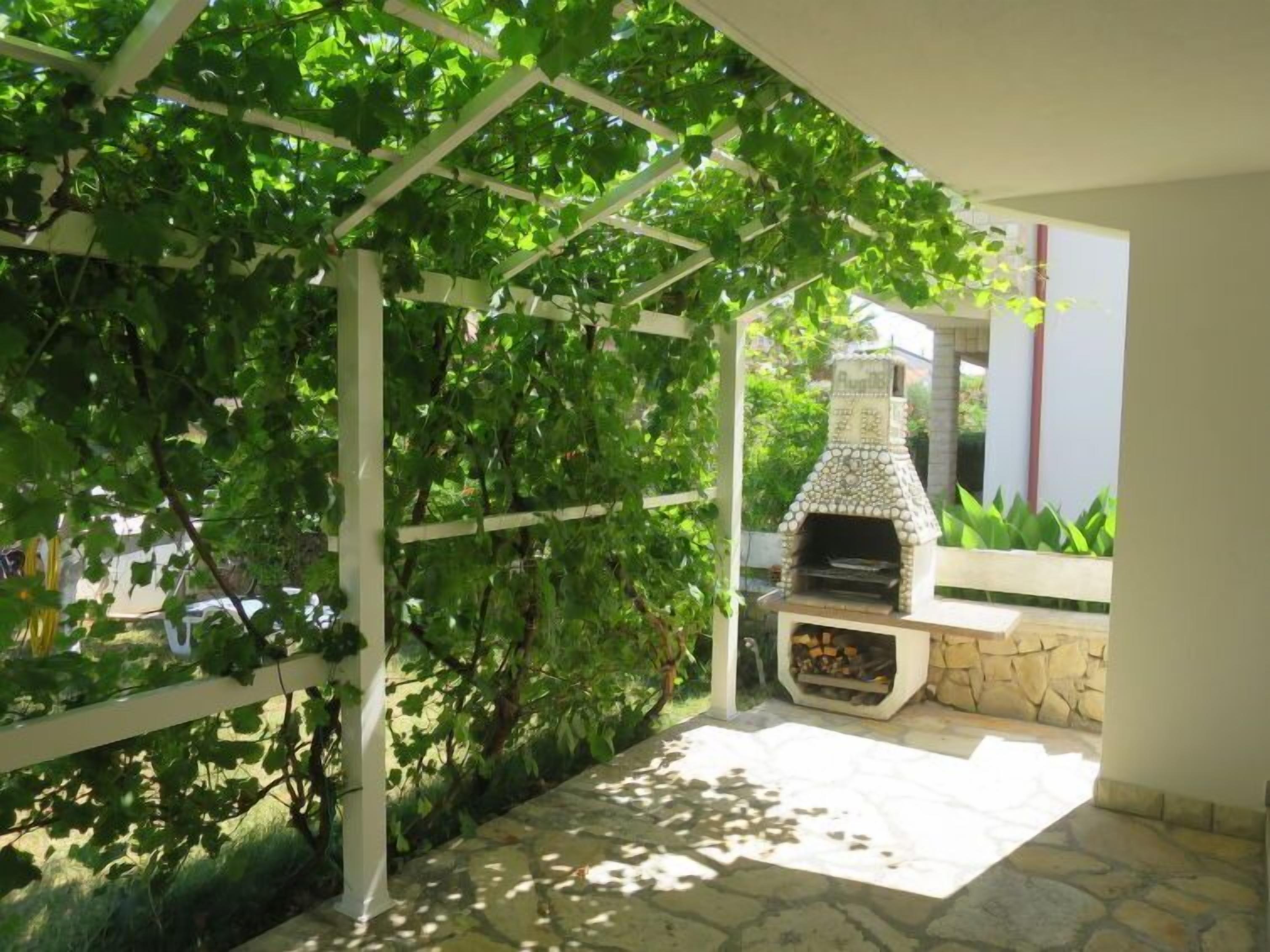 An outdoor patio with a stone barbecue grill, surrounded by greenery and a pergola structure.