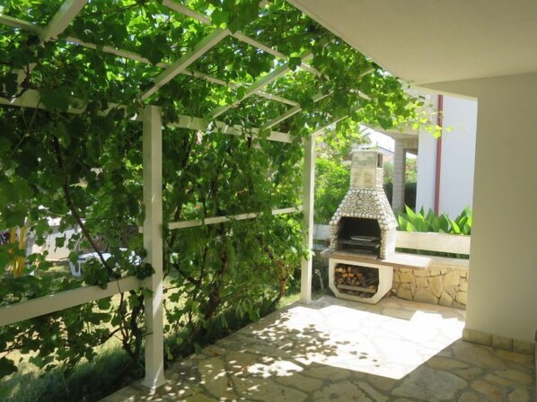 An outdoor patio with a stone barbecue grill, surrounded by greenery and a pergola structure.