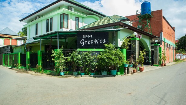 Un hôtel appelé « Hotel Greenia », avec une façade verte, une entrée couverte et des plantes en pot disposées autour de l’entrée.